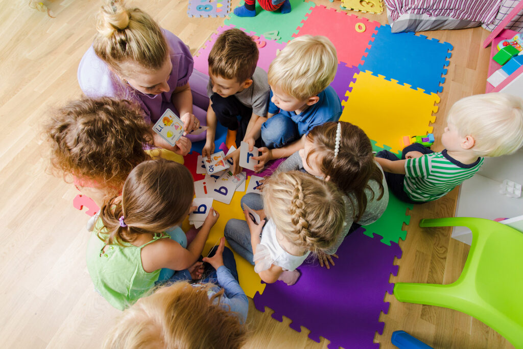 A group of children sit in a circle on colorful foam mats, playing with alphabet cards, while an adult supervises and helps.