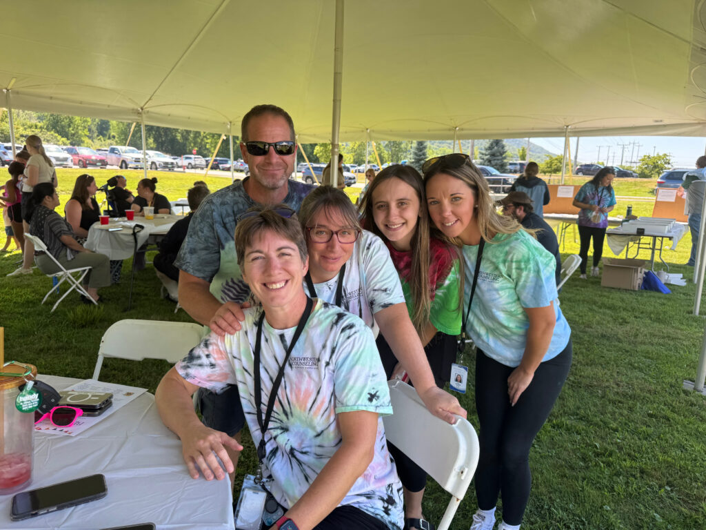 Five people wearing tie-dye shirts smile and pose together under a large outdoor tent at a grassy event. Other people are gathered at tables in the background. The atmosphere is casual and friendly.