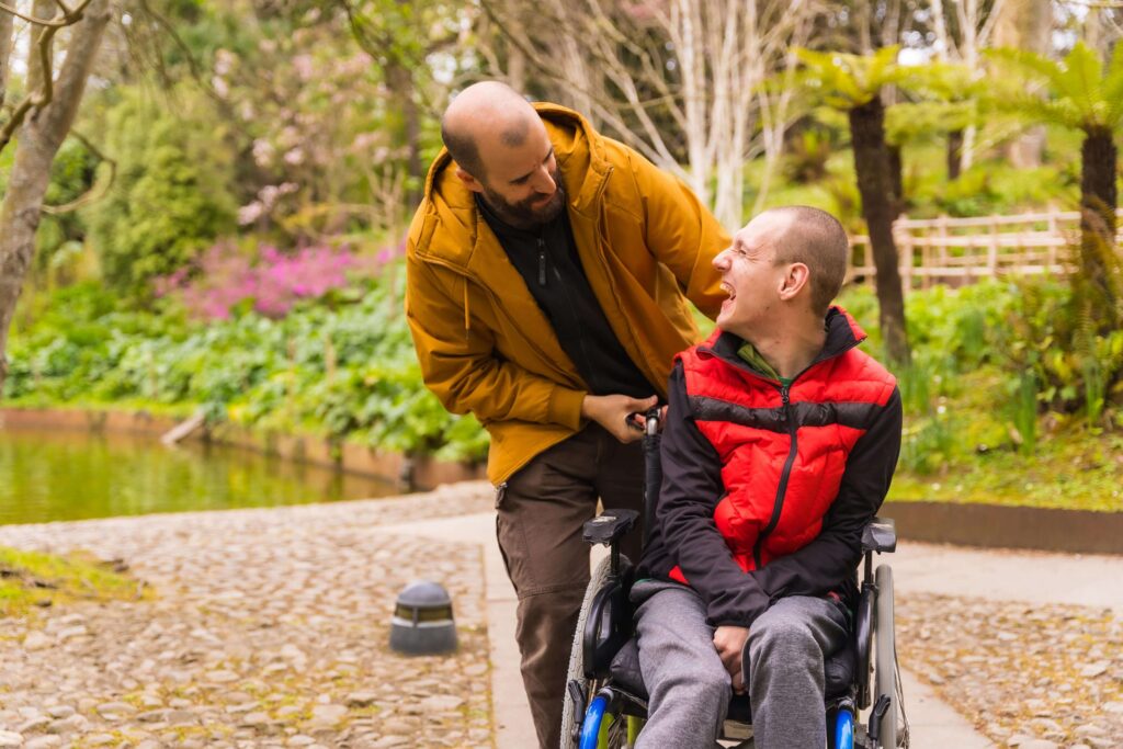 Two men are outdoors; one man in a red and black vest sits in a wheelchair, smiling and looking up at the man behind him, who is pushing the chair. They appear happy, surrounded by greenery in a park setting.