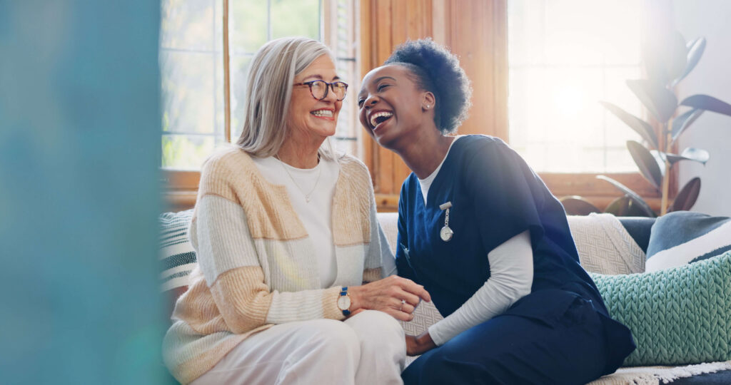An older woman and a caregiver sit close together on a couch, smiling and laughing, with sunlight streaming through large windows behind them.
