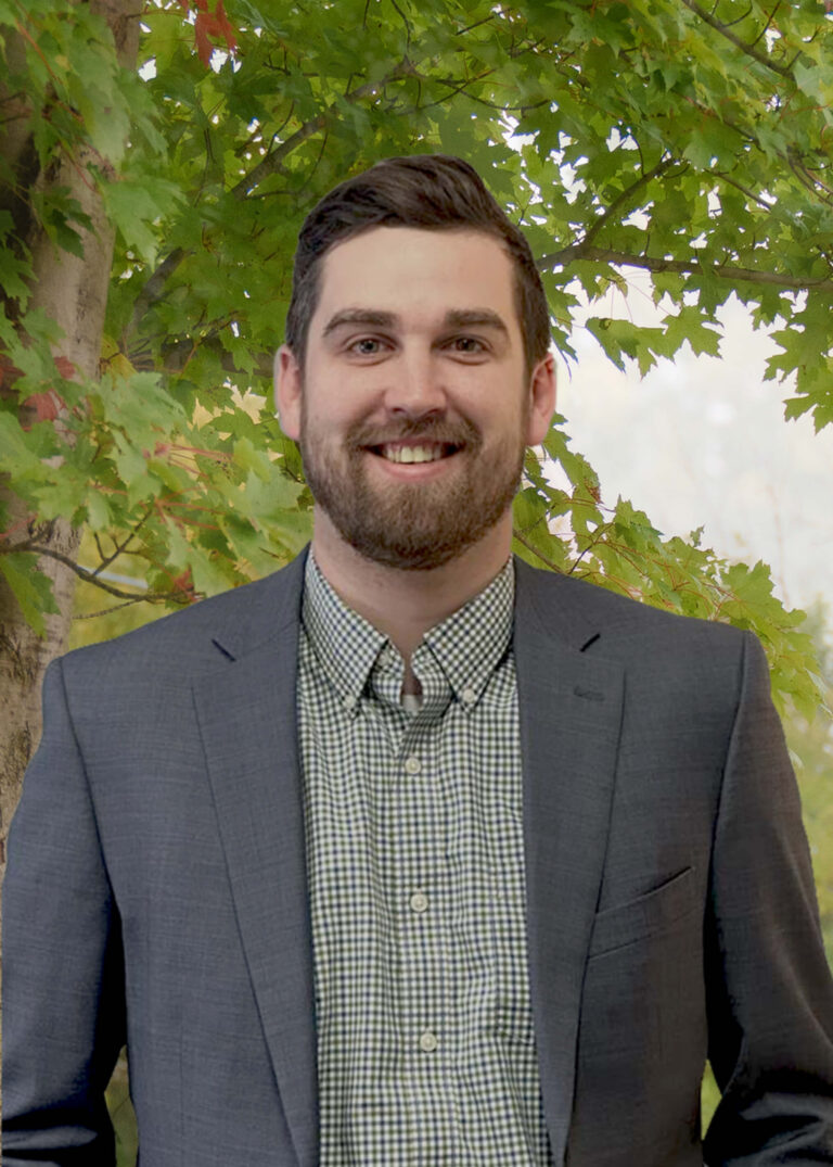 A man with short dark hair and a beard, wearing a checked shirt and gray blazer, stands smiling in front of green leafy trees outdoors.