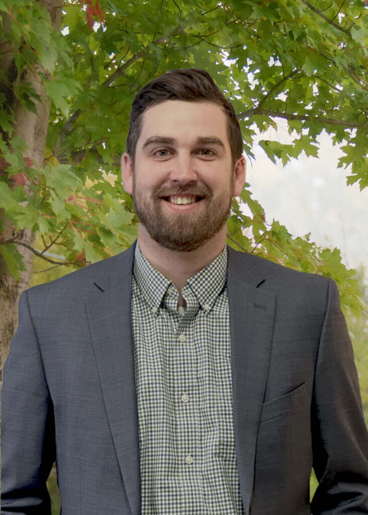 A man with brown hair and a beard, wearing a gray blazer over a green checkered shirt, stands smiling in front of leafy green trees.