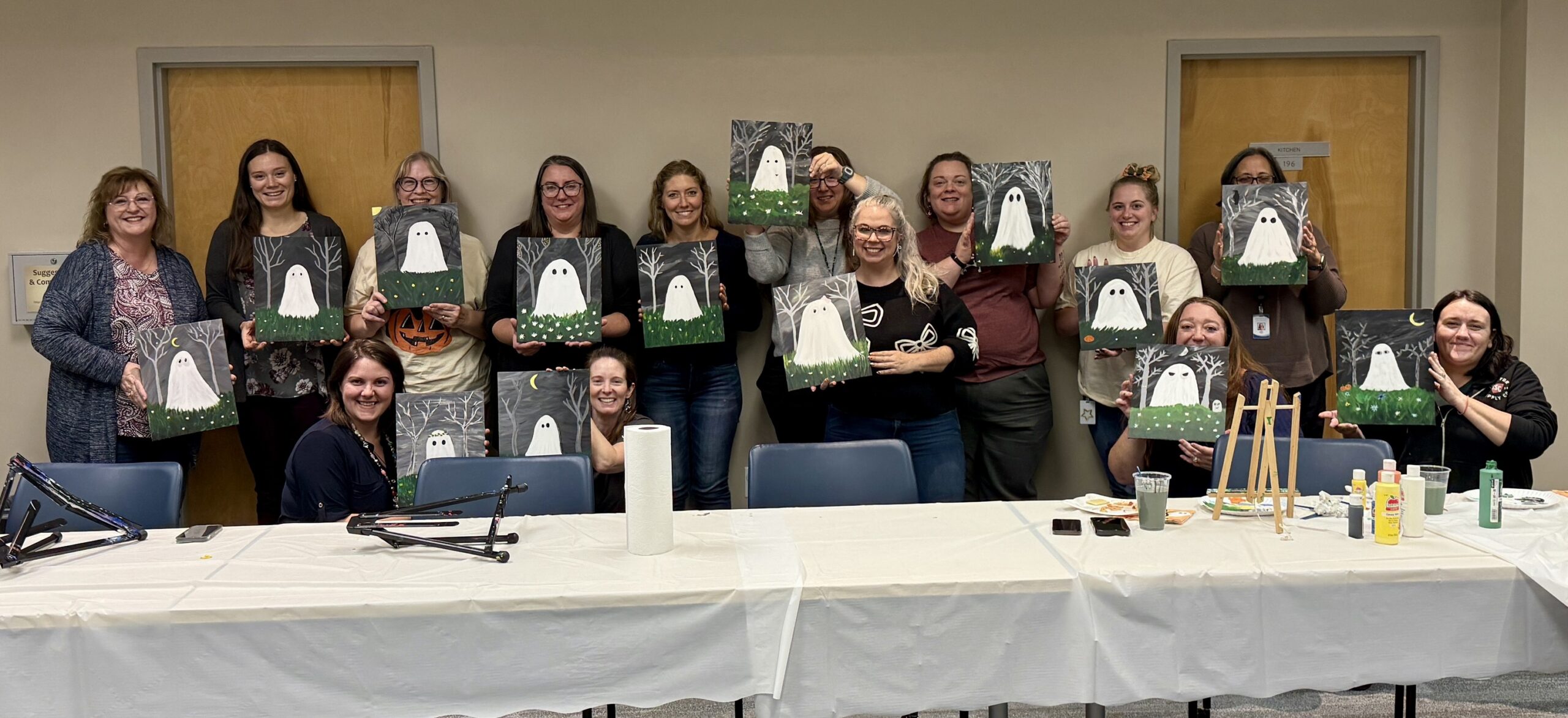 A group of people pose indoors, each holding a painting of a ghost in a grassy field. They are smiling and standing or sitting behind a table with art supplies and drinks.