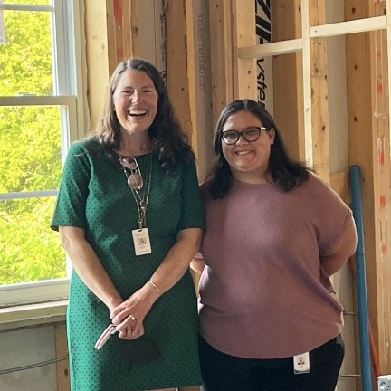 Two women stand and smile inside a building under construction, with exposed wooden beams and a window letting in natural light. Both wear ID badges around their necks.