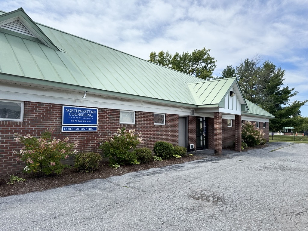 A brick building with a green metal roof and a sign reading Northwestern Counseling above some bushes; a paved parking area is in front, and trees are visible in the background.