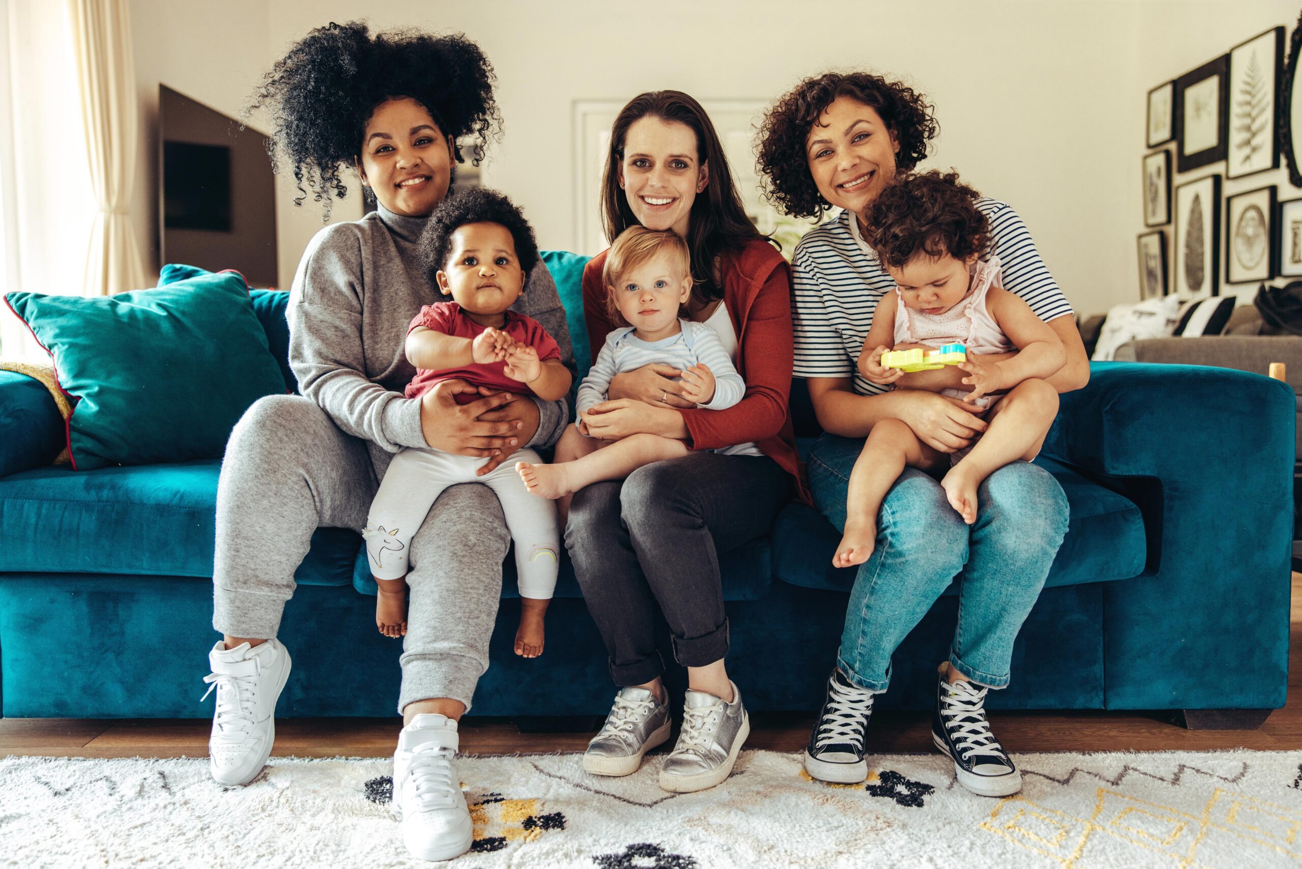 Three women sit on a blue couch, each holding a young child. They are smiling and looking at the camera in a cozy, well-lit living room decorated with framed art.
