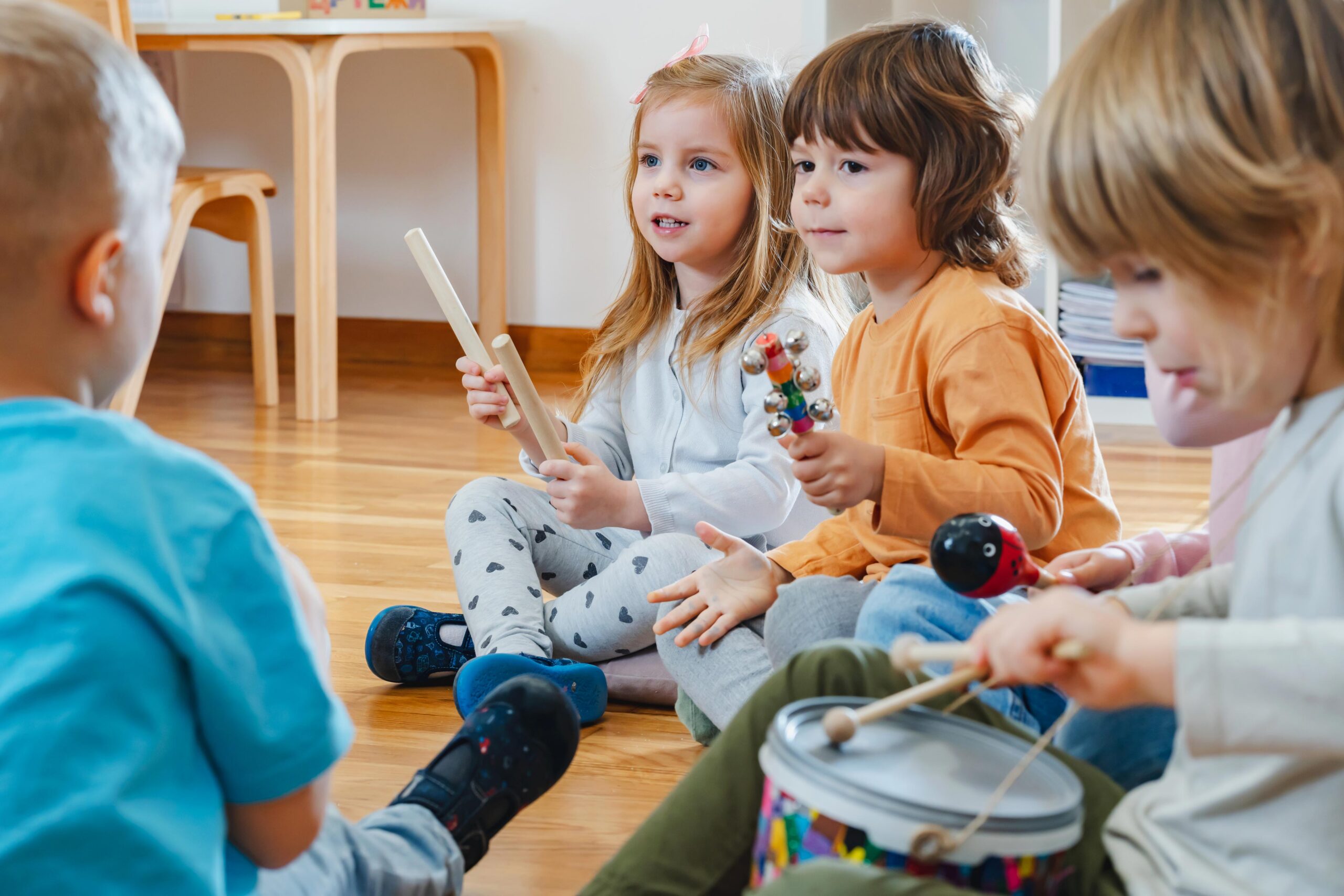 A group of young children sit in a circle on a wooden floor, playing musical instruments including a drum, maracas, and rhythm sticks, and smiling as they enjoy a music activity together.