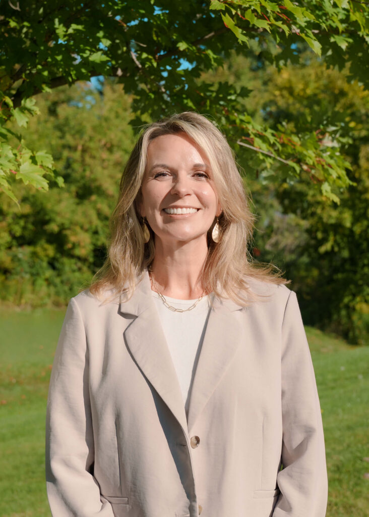 A woman with blonde hair wearing a beige blazer stands outside, smiling in front of green trees and grass on a sunny day.