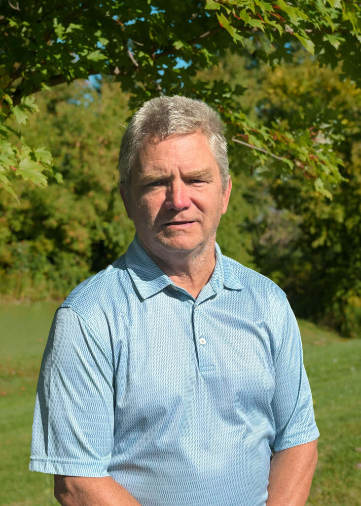 A man with short gray hair wearing a light blue polo shirt stands outdoors in front of green trees on a sunny day.