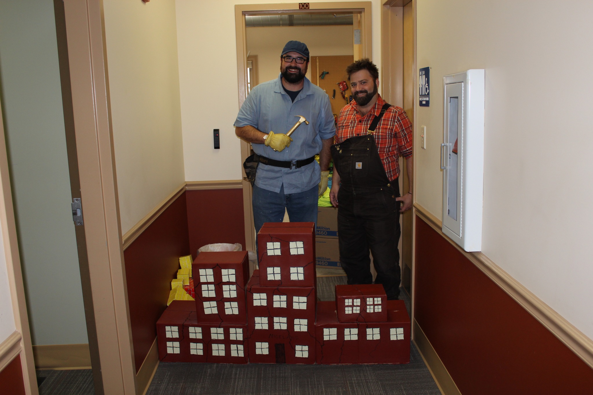 Two men in work clothes and gloves stand in a hallway, smiling, behind cardboard models of red buildings with white windows. One man holds a hammer and they appear to be posing for a themed event or costume.
