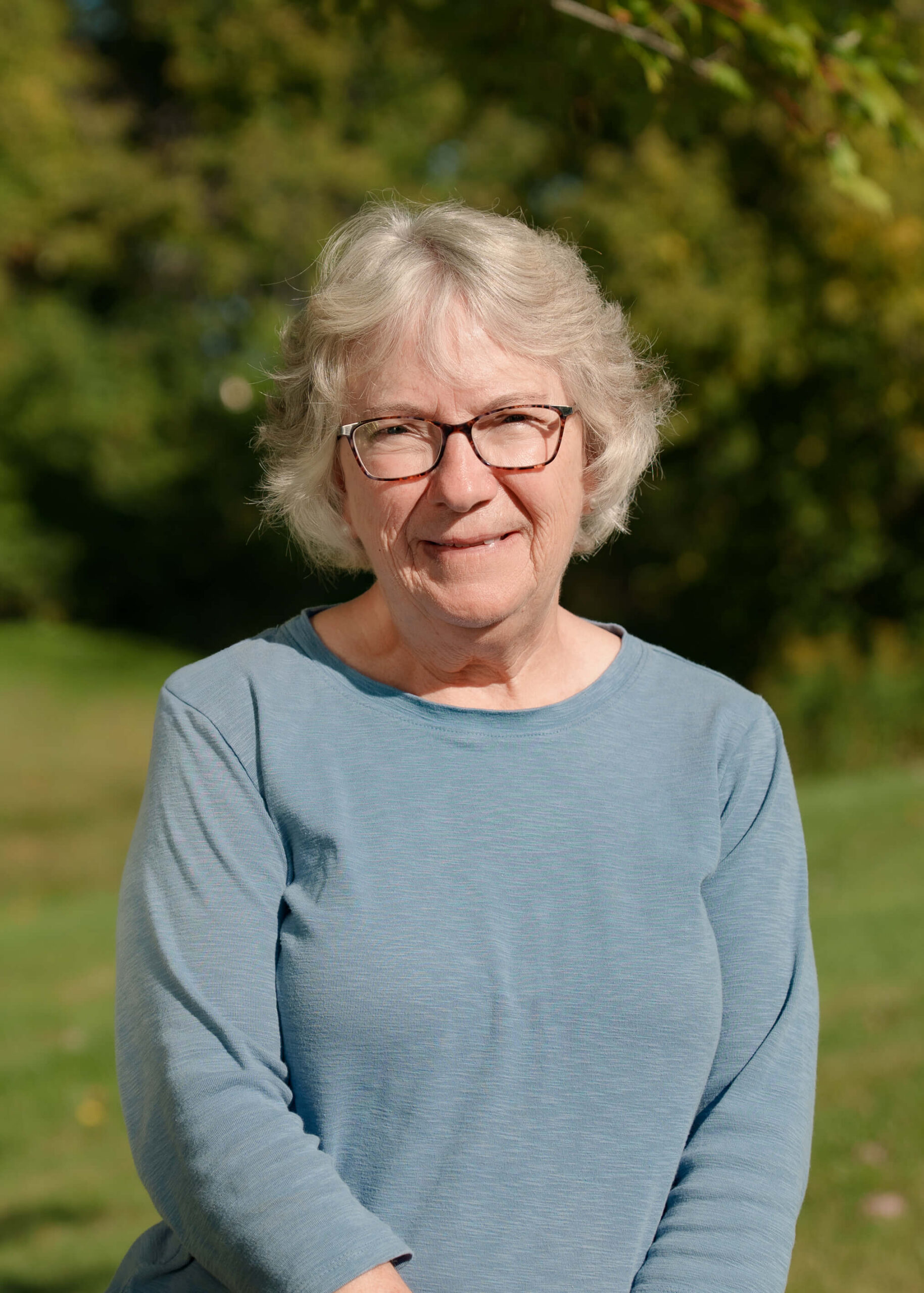 Older woman with short gray hair and glasses, wearing a blue long-sleeve shirt, smiling outdoors with trees and grass in the background.