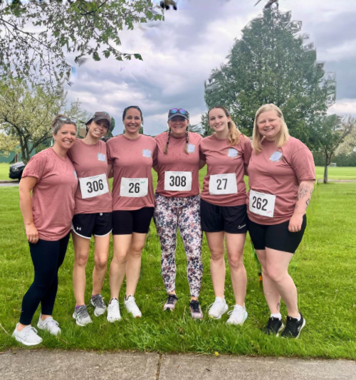 Six women in matching pink shirts and race bibs stand smiling together on grass, outdoors, under a cloudy sky. Trees are in the background and the group appears happy and ready for a race or event.