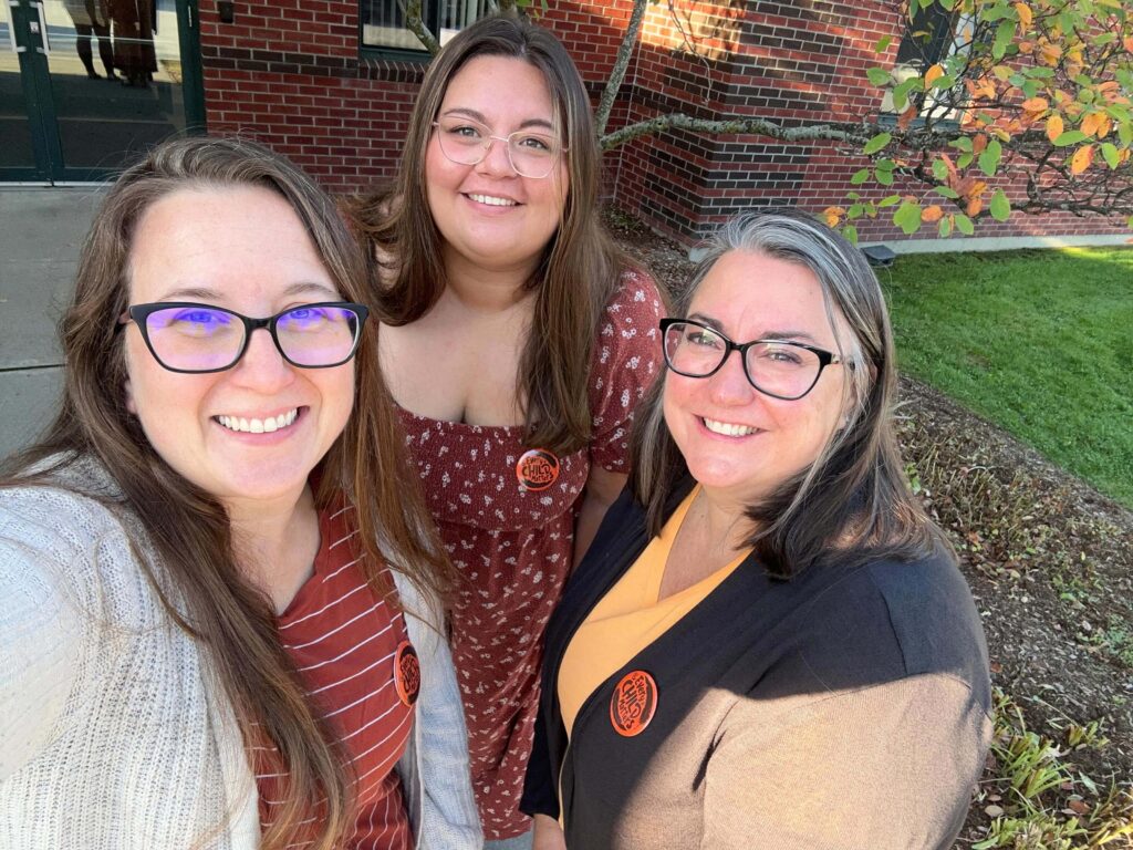 Three women wearing glasses and autumn-colored outfits smile outdoors beside a brick building and some fall foliage. All three have round orange stickers on their clothes.