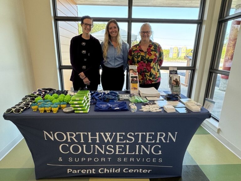 Three people stand behind a table with brochures, giveaways, and promotional items. The tablecloth reads Northwestern Counseling & Support Services Parent Child Center. Large windows are in the background.
