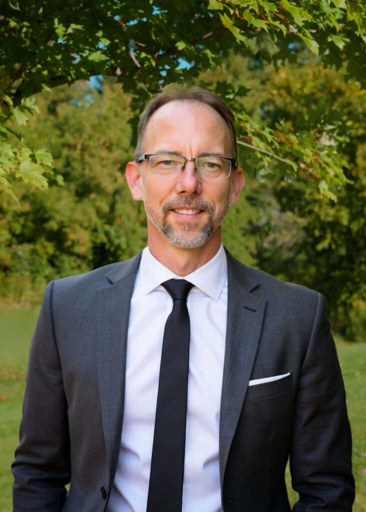 A man in a suit and tie stands outdoors in front of green trees, smiling at the camera. He has glasses, a short beard, and neatly styled hair.