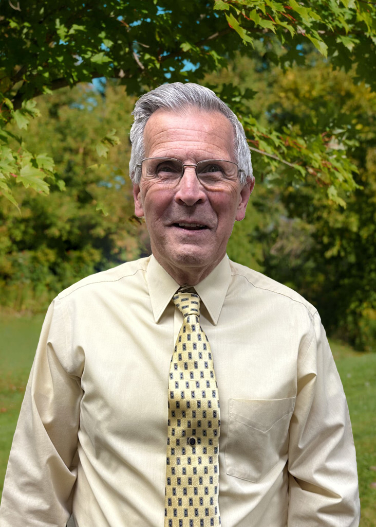 An older man with gray hair and glasses, wearing a light yellow shirt and patterned tie, stands outdoors in front of green trees on a sunny day.