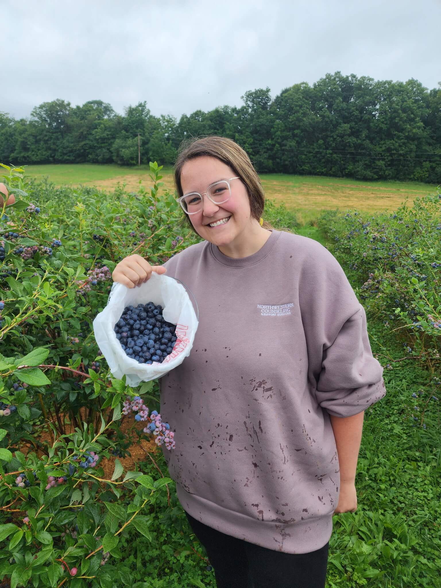 A person wearing a mauve sweatshirt and glasses smiles while holding a bag of freshly picked blueberries in a field, surrounded by green bushes and trees.