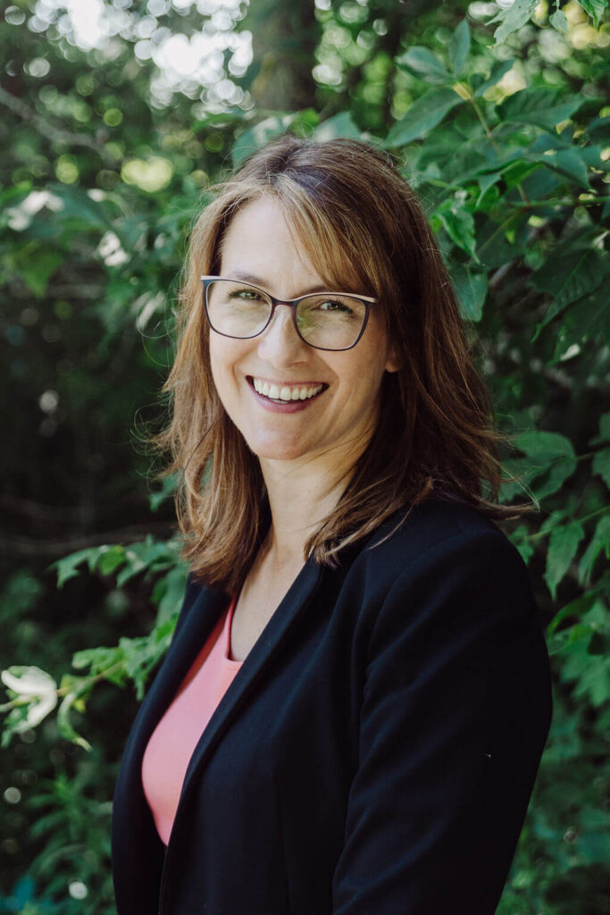 A woman with shoulder-length brown hair, glasses, and a black blazer over a pink top smiles outdoors with green foliage in the background.