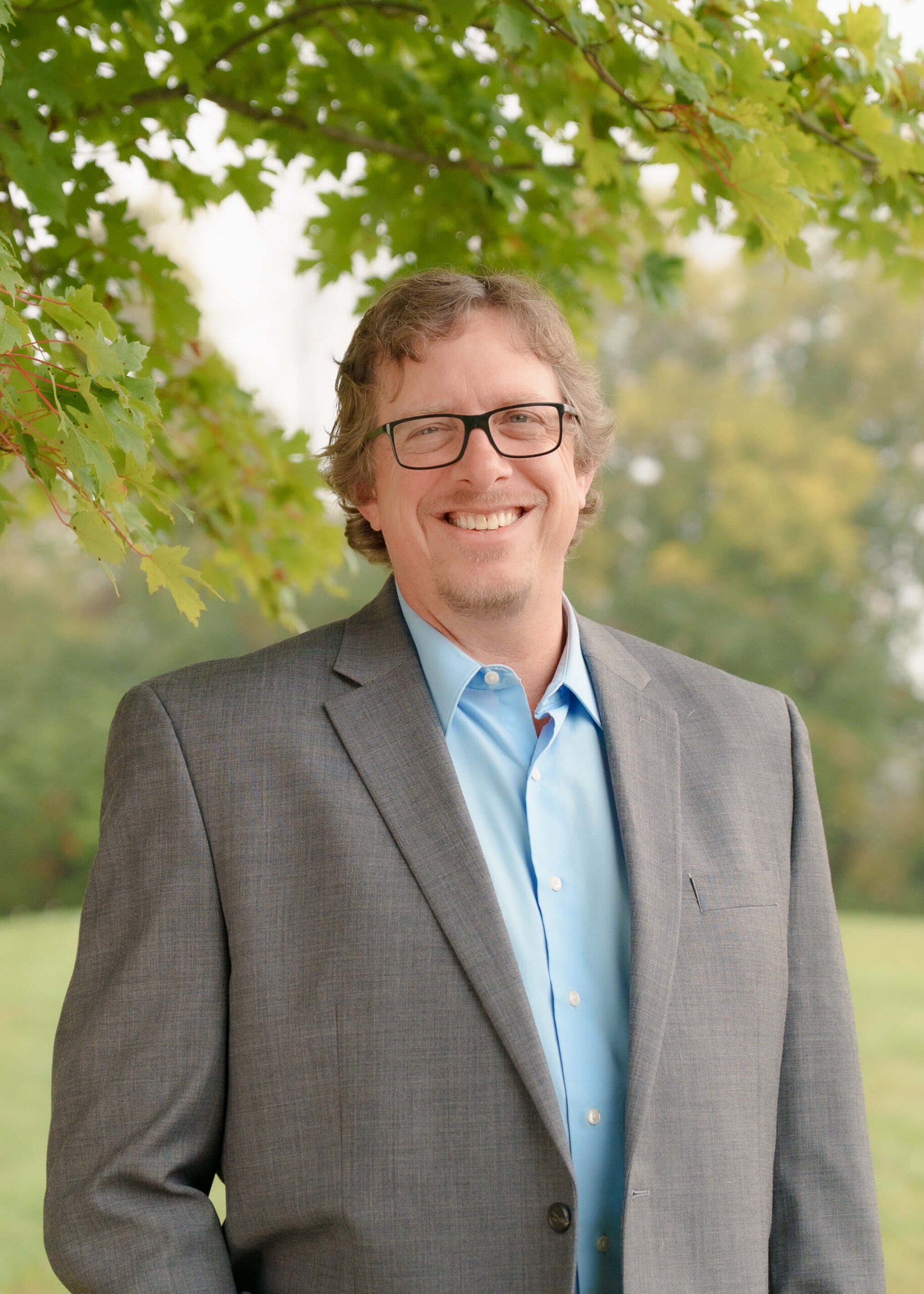 A man with glasses and wavy hair, wearing a gray suit and blue shirt, stands smiling outdoors beneath green tree branches with a blurred natural background.