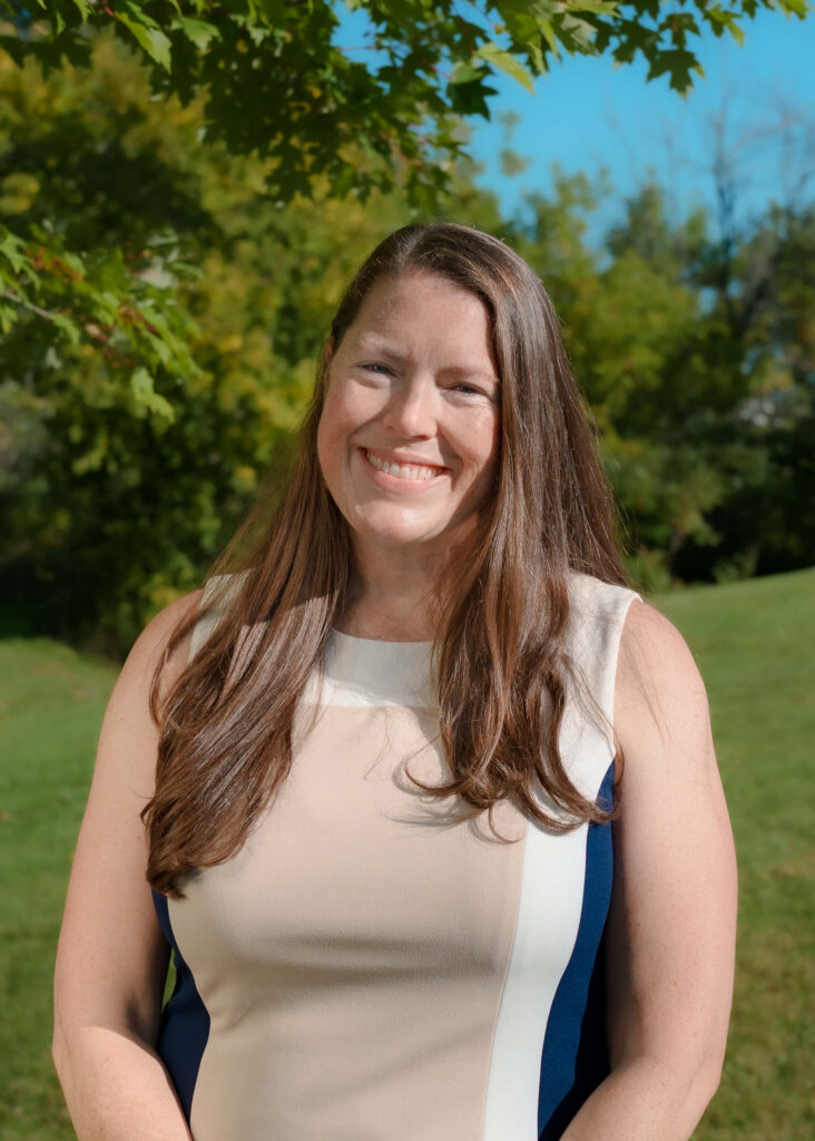 A woman with long brown hair wearing a sleeveless beige and white dress smiles outside, standing in front of green trees and grass under a sunny blue sky.