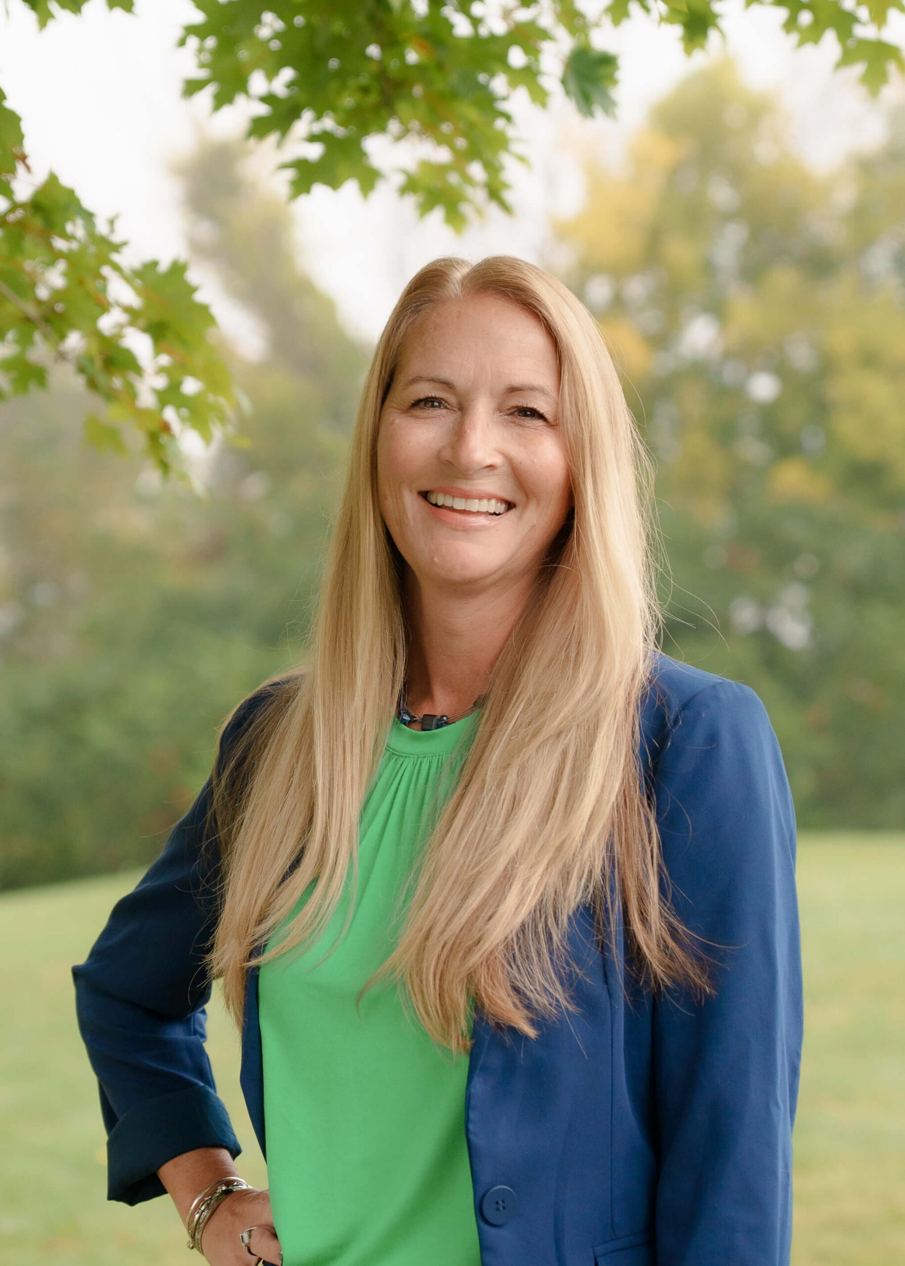 Woman with long blonde hair smiles outdoors, wearing a green top and blue blazer. Trees and greenery are in the background, giving a natural, relaxed setting.