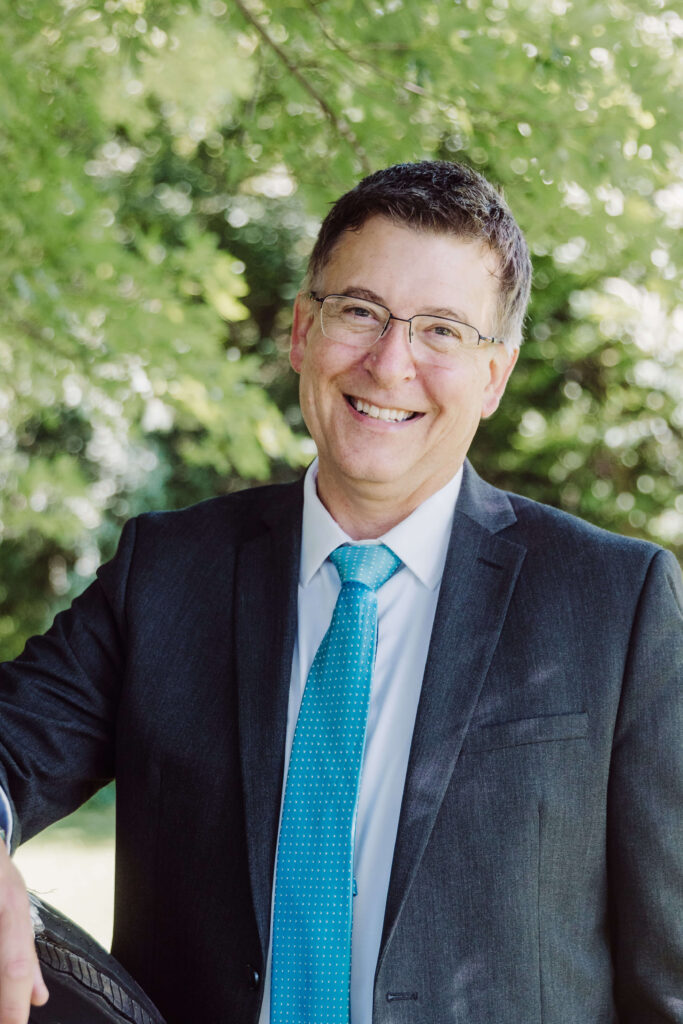 A man in a dark suit, white shirt, and blue tie smiles while standing outdoors with green foliage in the background.