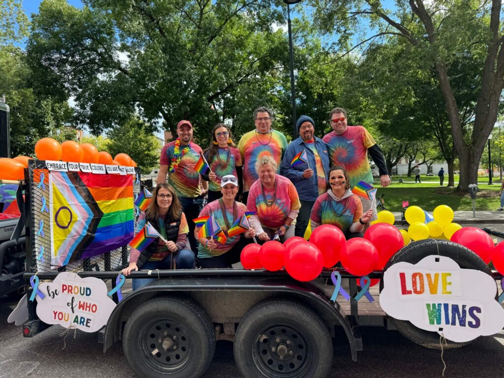A group of people in rainbow tie-dye shirts pose on a parade float decorated with Pride flags, red and yellow balloons, and signs reading LOVE WINS and Be PROUD OF WHO YOU ARE in a park setting.