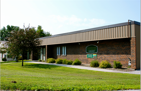 Single-story brick building with a flat roof, surrounded by green grass and shrubs. A tree stands by the entrance. A green sign with white text is mounted on the wall near the front door.