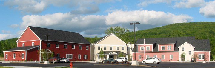 A large building complex with red and beige sections sits in front of a green, tree-covered hill under a partly cloudy sky. Several cars are parked in the lot in front of the buildings.