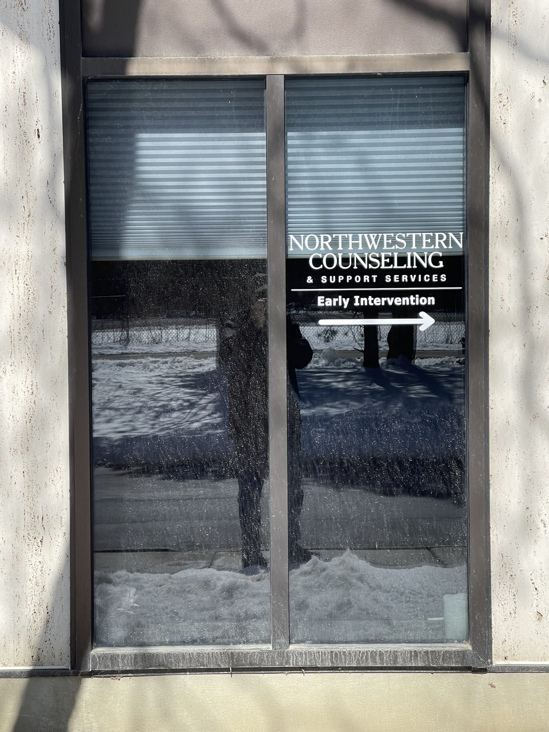 A window with a sign reading Northwestern Counseling & Support Services Early Intervention and an arrow pointing right. Snow is visible outside the window.