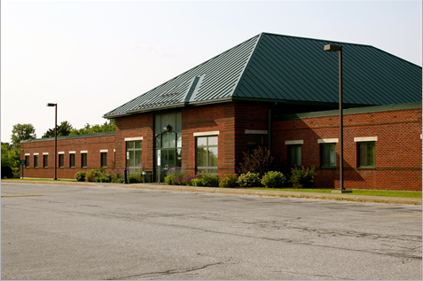 A single-story brick building with a green metal roof, several windows, and a main glass entrance, surrounded by landscaping and an empty parking lot.