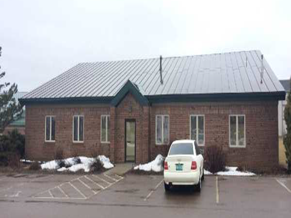 A single-story brick building with a metal roof, several windows, and a central entrance. A white car is parked in front, and patches of snow are visible on the ground.