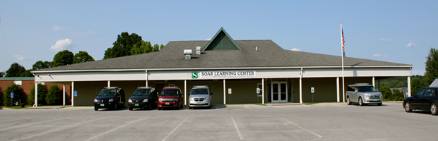 A single-story building labeled SOAR Learning Center with several cars parked in front, set on a clear day with trees in the background and an American flag on a pole to the right.