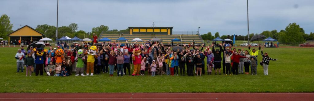 A large group of people, including several in colorful mascot costumes, stand together on a grassy field near a track, smiling and waving at the camera, with bleachers and tents in the background.