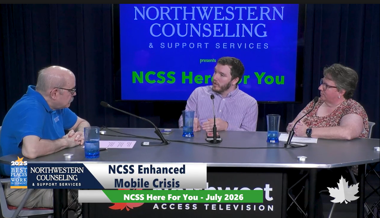 Three people sit at a table in a TV studio discussing NCSS Enhanced Mobile Crisis, with microphones and water glasses in front of them. A screen behind them reads NCSS Here for You.