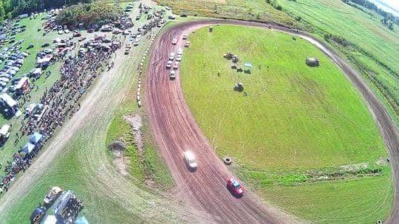 Aerial view of a dirt racetrack with several cars racing, surrounded by grassy fields. Spectators, cars, and tents are gathered on the left side of the image.