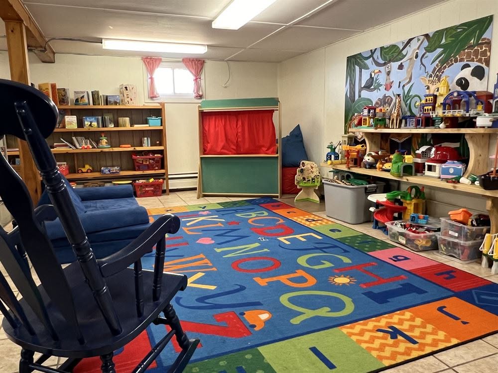 855331091_1988717015849074_4147680809239124038_n A playroom featuring a black rocking chair, shelves filled with toys and books, a small puppet theater, and an alphabet-themed area rug.